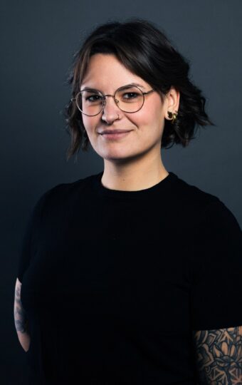 Anna Jennewein, mit kurzen dunklen Haaren, Brille und sichtbaren Armtattoos, steht in einem schwarzen Hemd vor einem dunklen Hintergrund und lächelt leicht.
