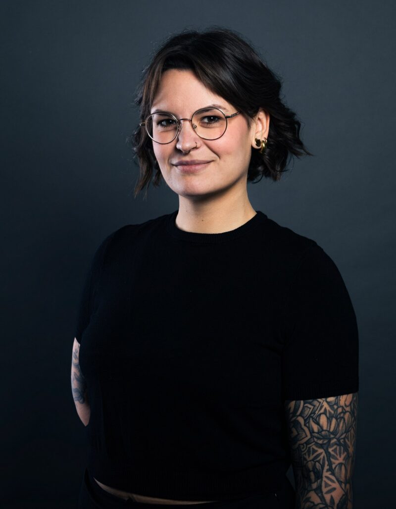 Anna Jennewein, mit kurzen dunklen Haaren, Brille und sichtbaren Armtattoos, steht in einem schwarzen Hemd vor einem dunklen Hintergrund und lächelt leicht.