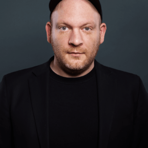 Marc Hellenthal, ein Mann mit hellem Teint, trägt eine schwarze Kappe, einen Blazer und ein Hemd, während er vor einem dunkelgrauen Hintergrund posiert. Er hat einen gestutzten Bart und blickt mit neutralem Blick direkt in die Kamera.