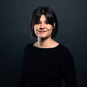 Lara Wollbold, eine Frau mit dunklem Haar und einem leichten Lächeln, steht vor einem dunkelgrauen Hintergrund und trägt ein schwarzes langärmeliges Oberteil.