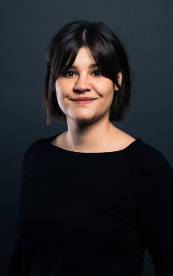 Lara Wollbold, eine Frau mit dunklem Haar und einem leichten Lächeln, steht vor einem dunkelgrauen Hintergrund und trägt ein schwarzes langärmeliges Oberteil.