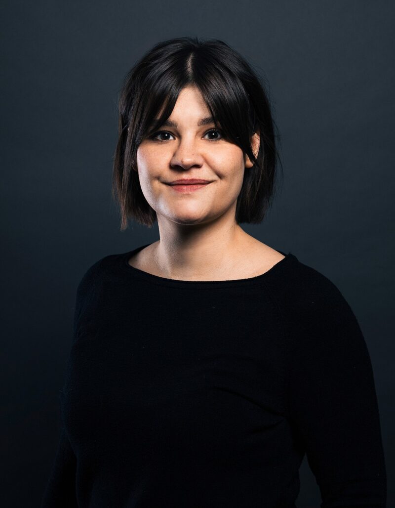 Lara Wollbold, eine Frau mit dunklem Haar und einem leichten Lächeln, steht vor einem dunkelgrauen Hintergrund und trägt ein schwarzes langärmeliges Oberteil.