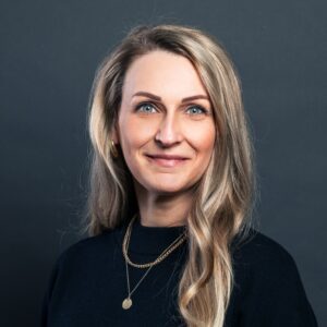 Eine Frau mit langen blonden Haaren, die ein schwarzes Oberteil und goldene Ketten trägt, steht vor einem dunkelgrauen Hintergrund und lächelt in die Kamera.