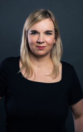 Sandra Bömer, mit glattem blondem Haar und einem schwarzen kurzärmeligen Oberteil, steht vor einem dunklen, schlichten Hintergrund und schaut selbstbewusst in die Kamera.