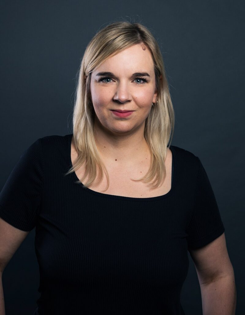 Sandra Bömer, mit glattem blondem Haar und einem schwarzen kurzärmeligen Oberteil, steht vor einem dunklen, schlichten Hintergrund und schaut selbstbewusst in die Kamera.