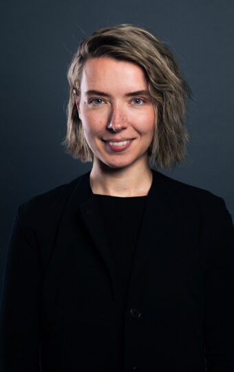 Eine Person mit kurzem, gewelltem blondem Haar, die eine schwarze Jacke über einem schwarzen Hemd trägt, steht vor einem dunklen, schlichten Hintergrund und lächelt in die Kamera.