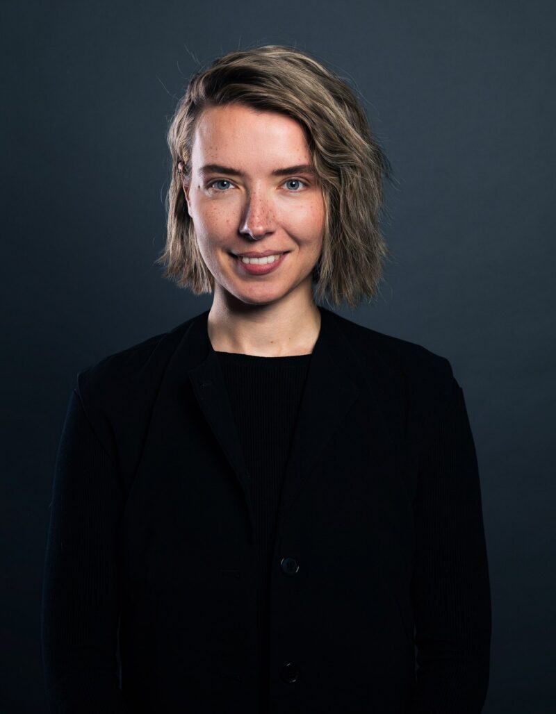 Eine Person mit kurzem, gewelltem blondem Haar, die eine schwarze Jacke über einem schwarzen Hemd trägt, steht vor einem dunklen, schlichten Hintergrund und lächelt in die Kamera.