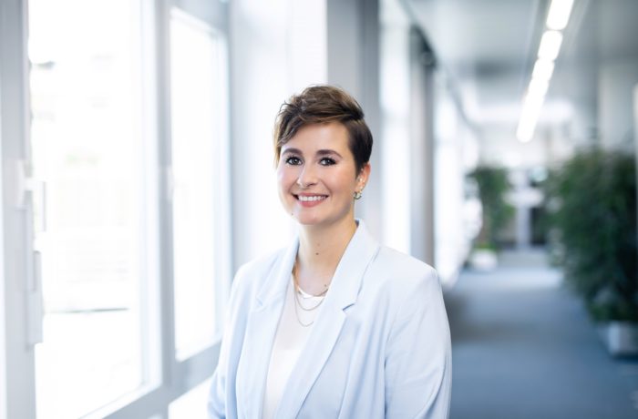 Eine Person mit kurzen braunen Haaren, die einen hellblauen Blazer und ein weißes Oberteil trägt, lächelt, während sie in einem hellen Flur mit Fenstern und Pflanzen im Hintergrund steht.