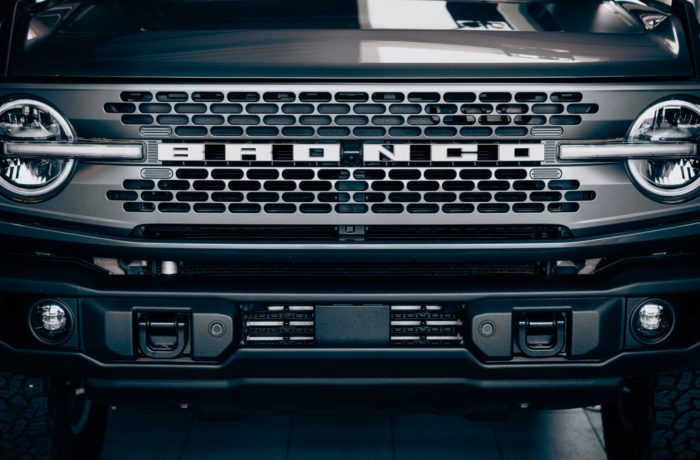 Nahaufnahme des Kühlergrills eines dunkel gefärbten Ford Bronco mit dem markanten BRONCO-Schriftzug, den runden Scheinwerfern und den robusten Stoßfängerdetails.