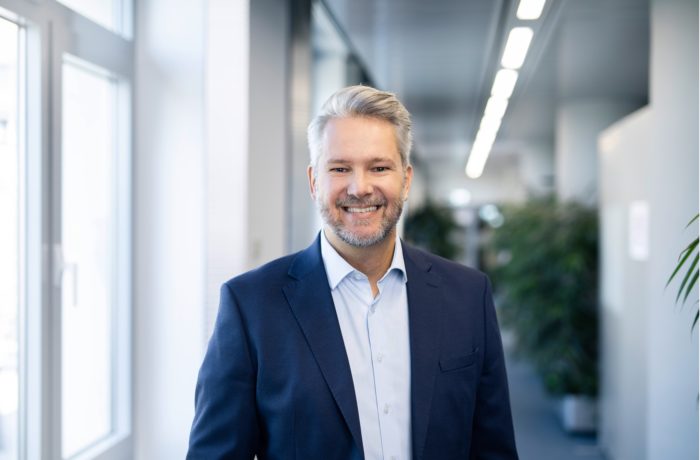 Ein lächelnder Mann mit grauem Haar und Bart, der einen blauen Anzug und ein hellblaues Hemd trägt, steht in einem modernen Büroflur mit großen Fenstern und Grünflächen im Hintergrund.