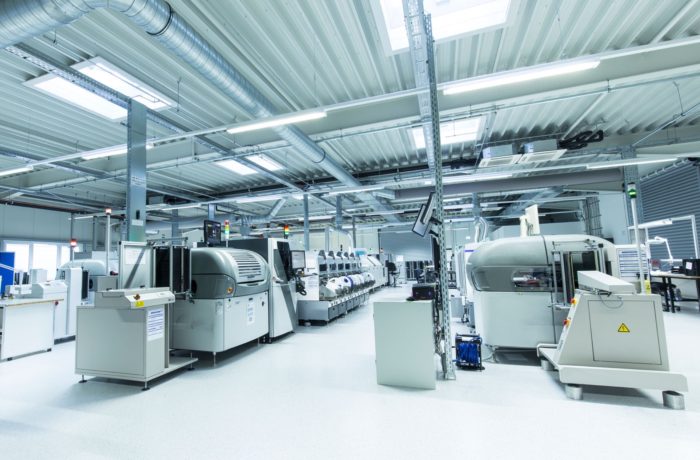 Eine geräumige, moderne Fabrikhalle mit großen Industriemaschinen, heller Deckenbeleuchtung und hellem Bodenbelag. Verschiedene automatisierte Geräte und Arbeitsstationen sind in geordneten Reihen angeordnet.