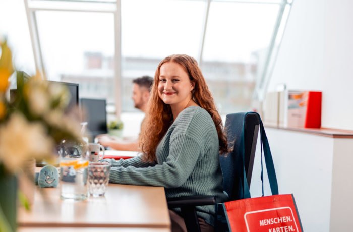 Eine Frau mit langen lockigen Haaren und einem grünen Pullover sitzt an einem Schreibtisch in einem hellen Büro und lächelt in die Kamera. Eine rote Tragetasche hängt an ihrem Stuhl, und im Hintergrund arbeitet eine weitere Person an einem Computer.