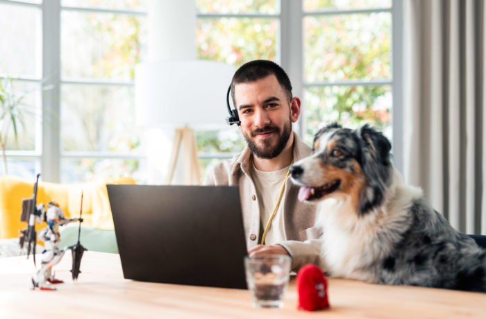 Ein Mann, der ein Headset trägt, lächelt, während er zu Hause an einem Laptop arbeitet. Ein Hund sitzt neben ihm am Tisch. Ein Glas Wasser, ein kleines rotes Spielzeug und ein Robotermodell stehen ebenfalls auf dem Tisch. Im Hintergrund sind helle Fenster zu sehen.