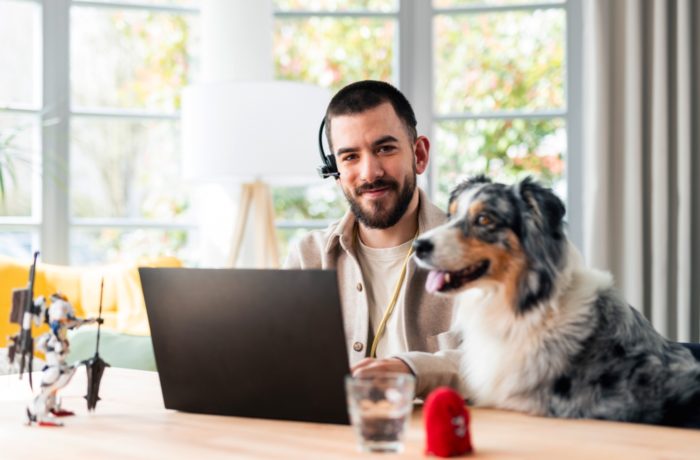 Ein Mann, der ein Headset trägt, sitzt an einem Schreibtisch mit einem Laptop und lächelt in die Kamera. Ein Hund sitzt neben ihm. Auf dem Tisch befinden sich ein Spielzeug, ein Glas Wasser und ein roter Gegenstand. Im Hintergrund sind helle Fenster und Grünflächen zu sehen.