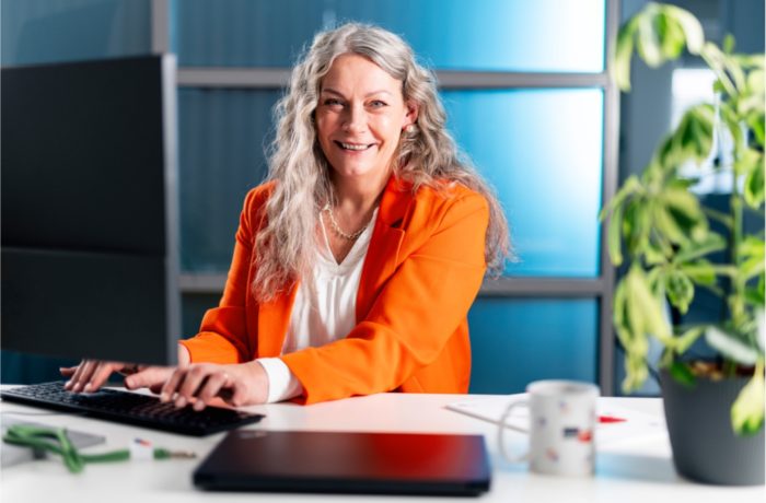 Eine lächelnde Frau mit langen grauen Haaren, die einen orangefarbenen Blazer trägt, sitzt an einem Schreibtisch und tippt auf einer Tastatur. Ein Computermonitor, eine Pflanze und ein Kaffeebecher stehen auf dem Schreibtisch in einer hellen Büroumgebung.