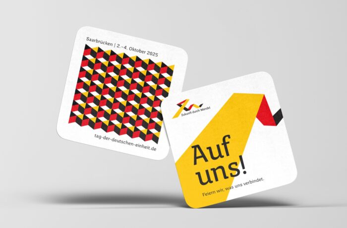 Zwei quadratische Untersetzer: einer mit einem geometrischen rot-gelb-schwarzen Würfelmuster und Veranstaltungshinweisen für Saarbrücken, 2. bis 4. Oktober 2025; der andere mit "Auf uns!" und einem stilisierten gelb-rot-schwarzen Bandmuster.