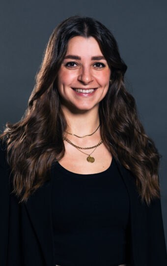 Carina Sutor, eine Frau mit langen braunen Haaren, die ein schwarzes Oberteil, eine schwarze Jacke und goldene Halsketten trägt, lächelt vor einem schlichten dunkelgrauen Hintergrund.