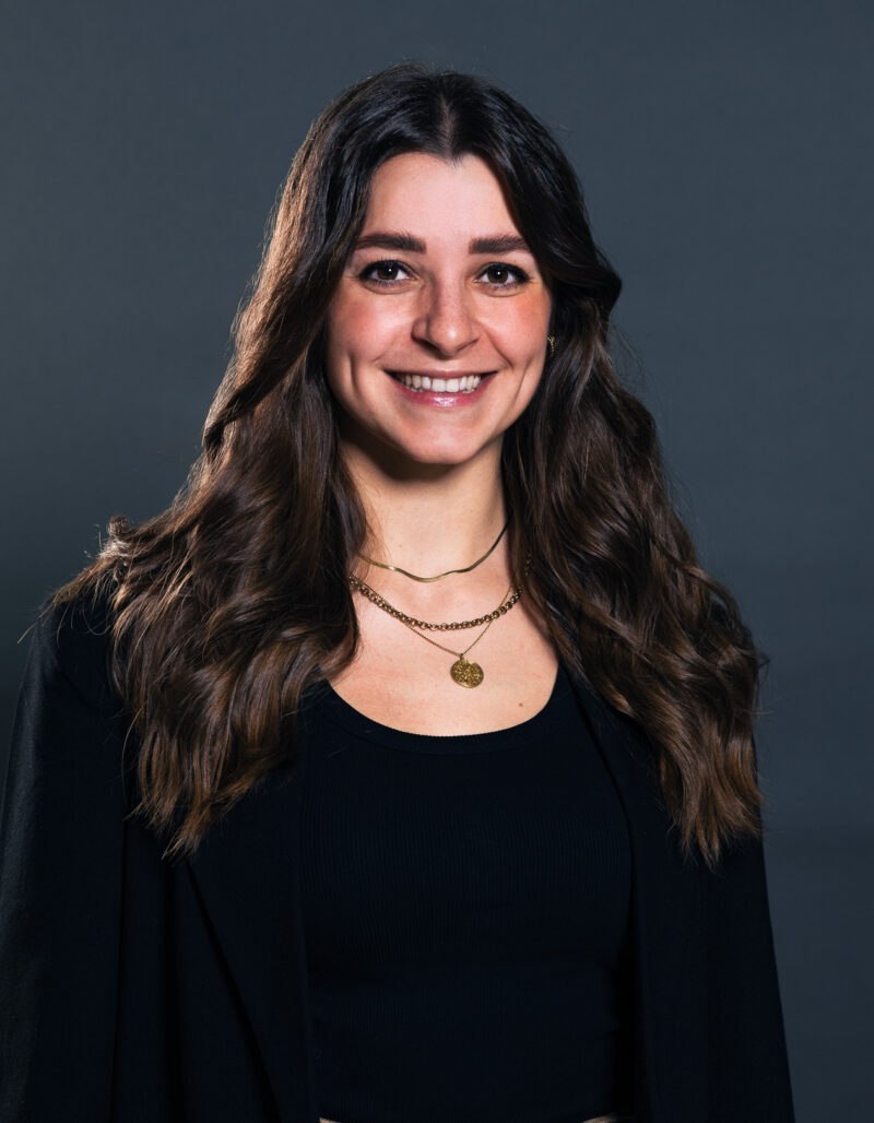 Carina Sutor, eine Frau mit langen braunen Haaren, die ein schwarzes Oberteil, eine schwarze Jacke und goldene Halsketten trägt, lächelt vor einem schlichten dunkelgrauen Hintergrund.