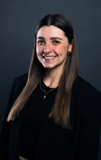 Michelle Elbert, eine junge Frau mit langen braunen Haaren, die einen schwarzen Blazer und goldene Halsketten trägt, lächelt warm vor einem dunklen, schlichten Hintergrund.