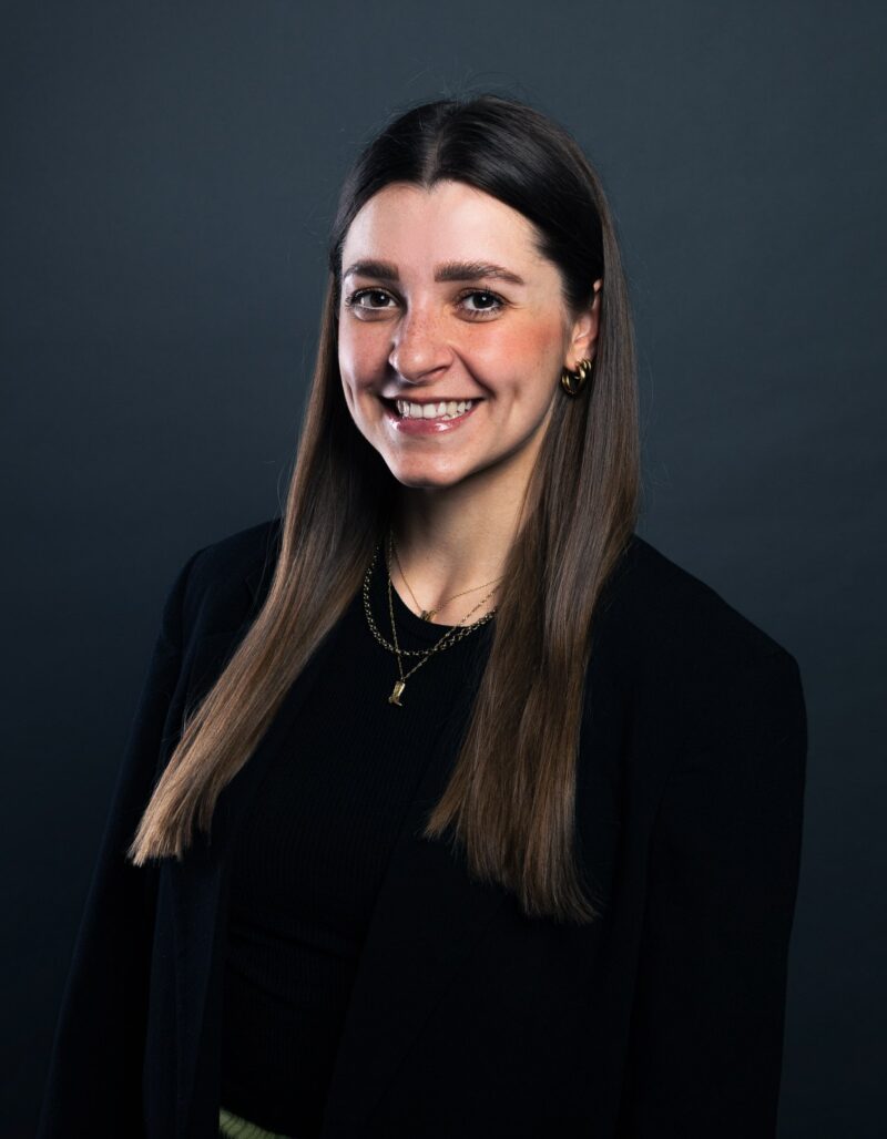 Michelle Elbert, eine junge Frau mit langen braunen Haaren, die einen schwarzen Blazer und goldene Halsketten trägt, lächelt warm vor einem dunklen, schlichten Hintergrund.