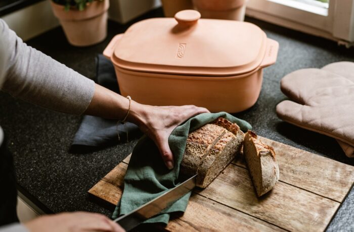 Während einer Event-Kampagne zum Tag der deutschen Einheit schneidet eine Person einen Laib Brot auf einem hölzernen Schneidebrett, das sie mit einem grünen Tuch abdeckt. Eine Terrakotta-Backform und Ofenhandschuhe liegen auf der schwarzen Arbeitsplatte daneben. Während einer Event-Kampagne zum Tag der deutschen Einheit schneidet eine Person einen Laib Brot auf einem hölzernen Schneidebrett, das sie mit einem grünen Tuch abdeckt. Eine Terrakotta-Backform und Ofenhandschuhe liegen auf der schwarzen Arbeitsplatte daneben.