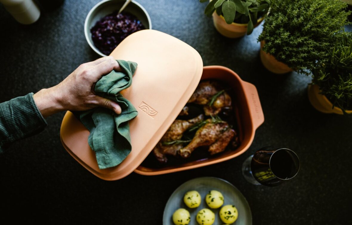 Eine Hand hebt den Deckel von einer Terrakotta-Backform mit gebratenen Hähnchenkeulen - perfekt für eine Event-Kampagne zum Tag der deutschen Einheit. Kräuter in Töpfen, gelbe Kartoffeln, rote Soße und ein Glas Rotwein stehen daneben auf einer dunklen Fläche.