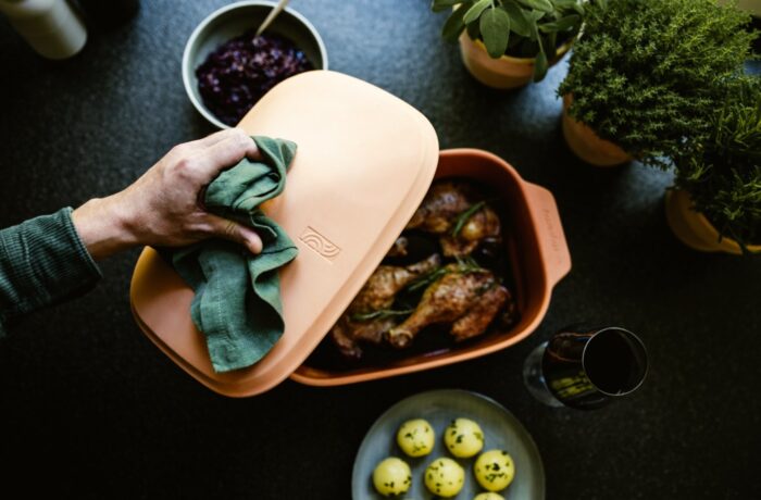 Eine Hand hebt den Deckel von einer Terrakotta-Backform mit gebratenen Hähnchenkeulen - perfekt für eine Event-Kampagne zum Tag der deutschen Einheit. Kräuter in Töpfen, gelbe Kartoffeln, rote Soße und ein Glas Rotwein stehen daneben auf einer dunklen Fläche. Eine Hand hebt den Deckel von einer Terrakotta-Backform mit gebratenen Hähnchenkeulen - perfekt für eine Event-Kampagne zum Tag der deutschen Einheit. Kräuter in Töpfen, gelbe Kartoffeln, rote Soße und ein Glas Rotwein stehen daneben auf einer dunklen Fläche.