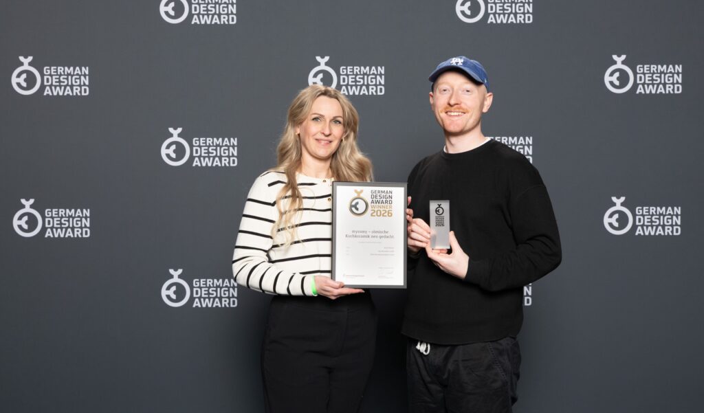 Zwei Personen stehen lächelnd vor einer Kulisse des German Design Award. Der eine hält eine gerahmte Urkunde in der Hand, der andere eine Trophäe. Beide feiern ihren Erfolg in Brand Identity für Kochgeschirr Marke.