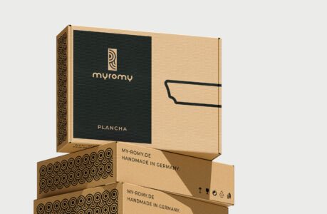 Ein Stapel von drei Pappschachteln mit myromy-Branding und dekorativen Mustern; die oberste Schachtel zeigt das Wort PLANCHA und eine minimalistische Pfannenillustration. Perfekt für eine Event-Kampagne zum Tag der deutschen Einheit; beschriftet mit HANDMADE IN GERMANY.