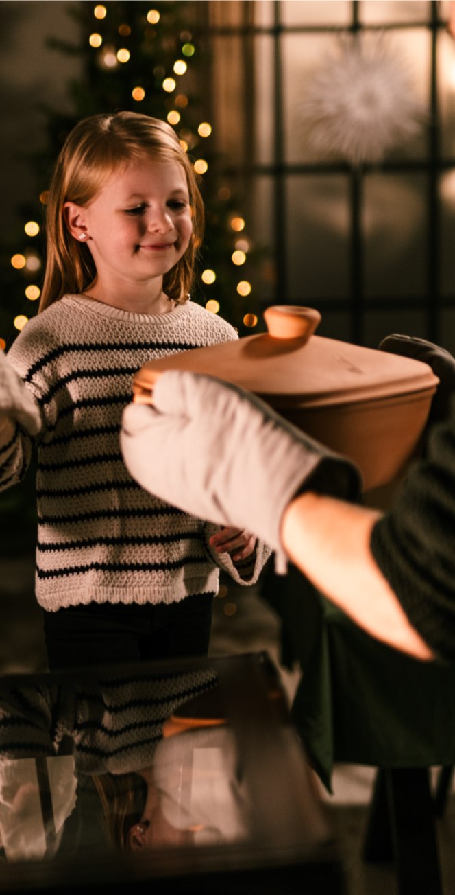 Ein junges Mädchen lächelt, als es von einem Mann mit Ofenhandschuhen einen gedeckten Teller erhält, während im Hintergrund ein geschmückter Weihnachtsbaum und festliche Lichter bei einer Event-Kampagne zum Tag der deutschen Einheit stehen.